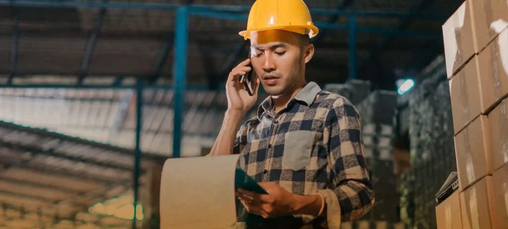 Warehouse worker in safety helmet multitasking with clipboard and phone, surrounded by stacked boxes and shelves.